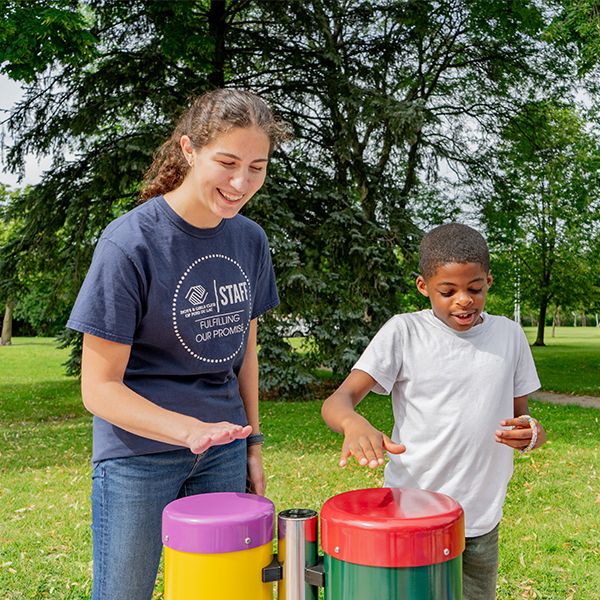 an older girl playing outdoor drums in a music park along with a young black boy