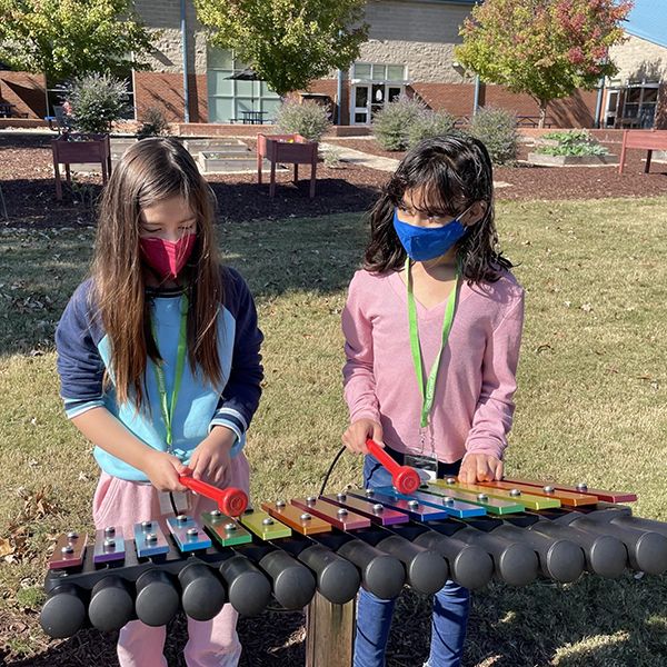 Two young girls wearing face masks playing outdoor musical instruments in their school playground