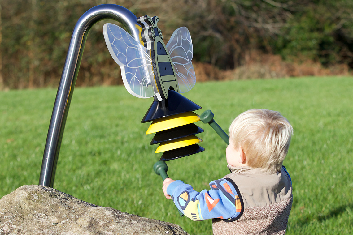 A child with blonde hair in colorful clothing plays with a bee-shaped musical instrument outdoors. The sunny setting features green grass and trees.