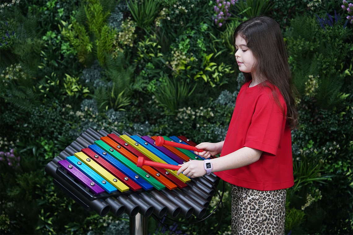 an outdoor xylophone with rainbow colored notes on a stainless steel leg with a young girl playing with red mallets and a green leafy background
