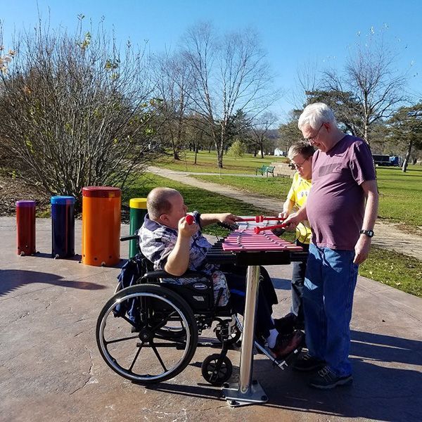 three people including a man in a wheelchair playing a duo outdoor xylophone in st feriole memorial gardens music park