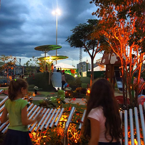 two girls playing a silver outdoor xylophone in a playground at night with LED lights in the plants and trees 