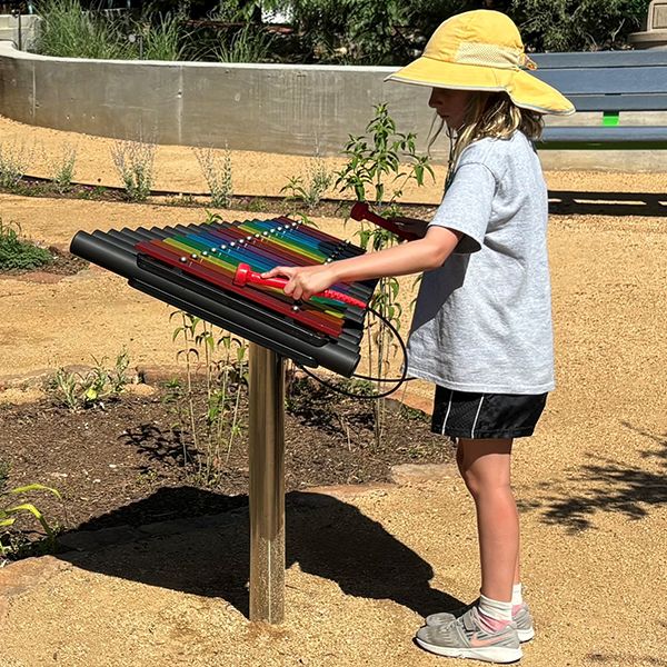 a young girl wearing a large brimmed hat and shorts playing an outdoor xylophone in a music playground