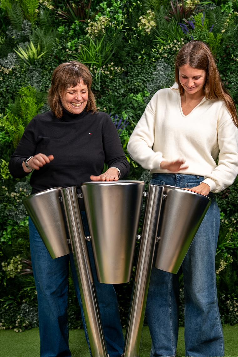 Two people playing outdoor stainless steel drums with their hands, surrounded by greenery. The scene conveys a joyful, musical interaction.