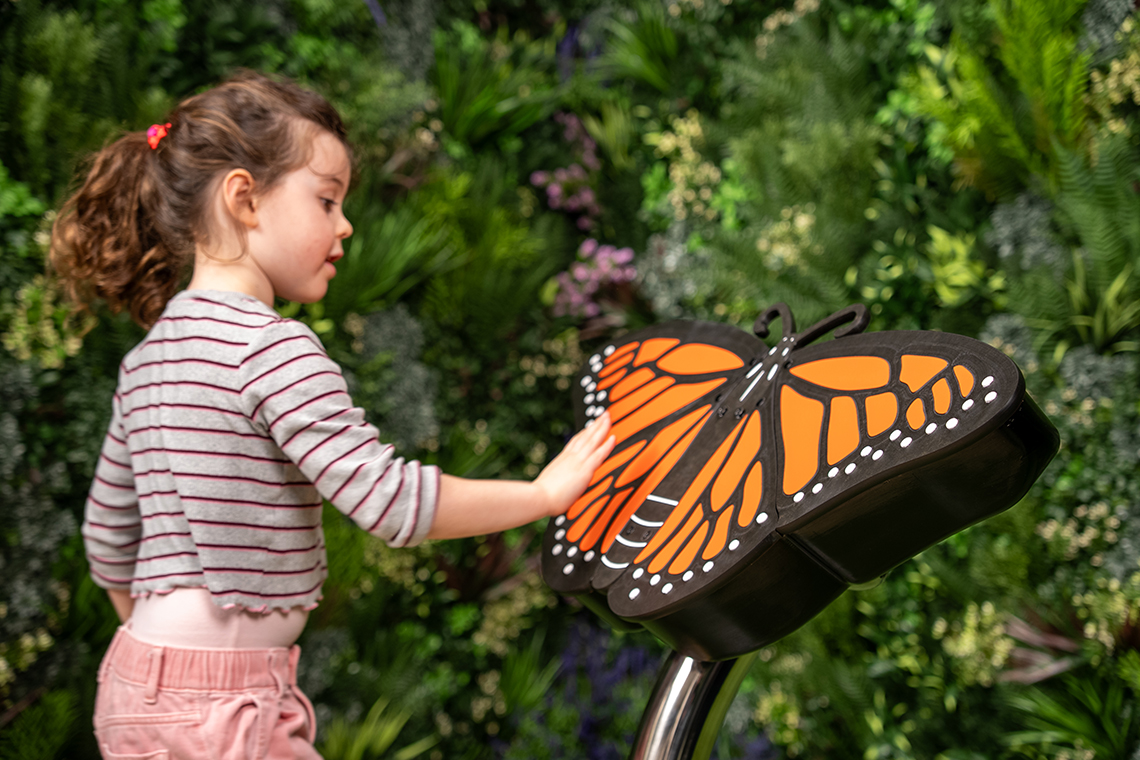 A child interacts using their hands with a large monarch butterfly musical sculpture against a vibrant green plant wall
