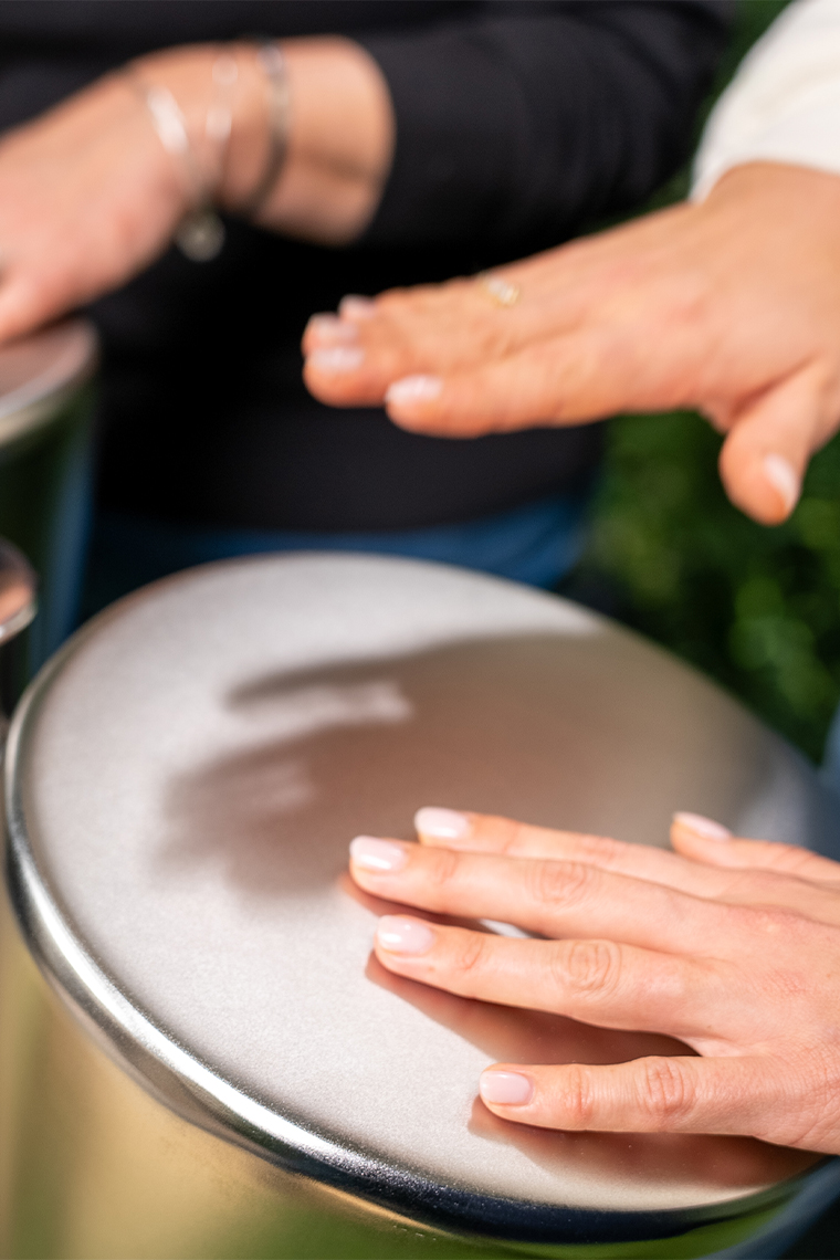 Close-up of two people playing outdoor stainless steel drums with their hands, surrounded by greenery. The scene conveys a joyful, musical interaction.