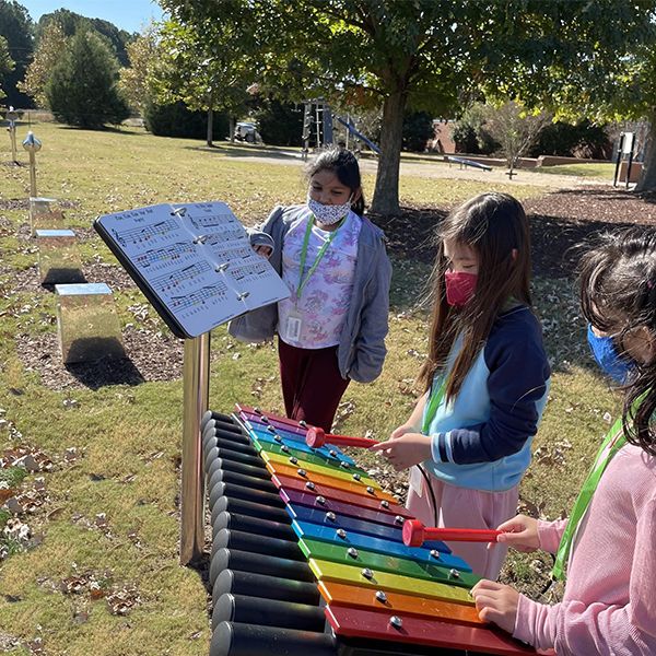 Three young girls wearing face masks playing outdoor musical instruments in their school playground