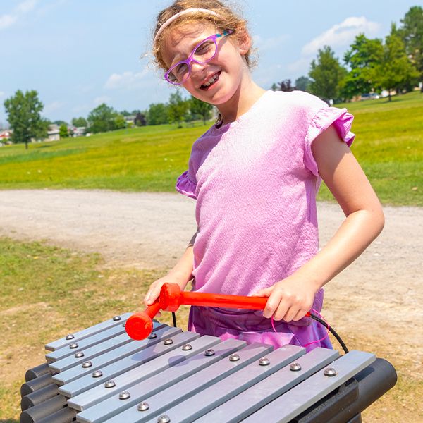 A red headed girl in glasses smiling playing an outdoor xylophone with red mallets