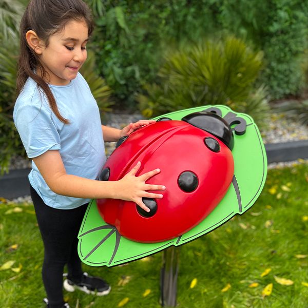 A young girl touching a large, red ladybug musical sculpture on a green leaf in a garden. She smiles with delight, surrounded by lush greenery.