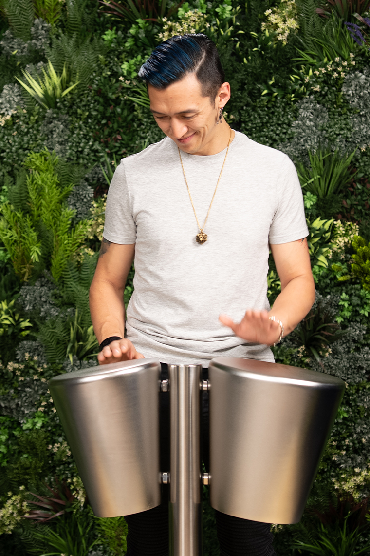 A person in a gray shirt energetically plays a pair of large stainless steel outdoor drums. They are surrounded by a lush vertical garden, exuding a lively and joyous vibe.