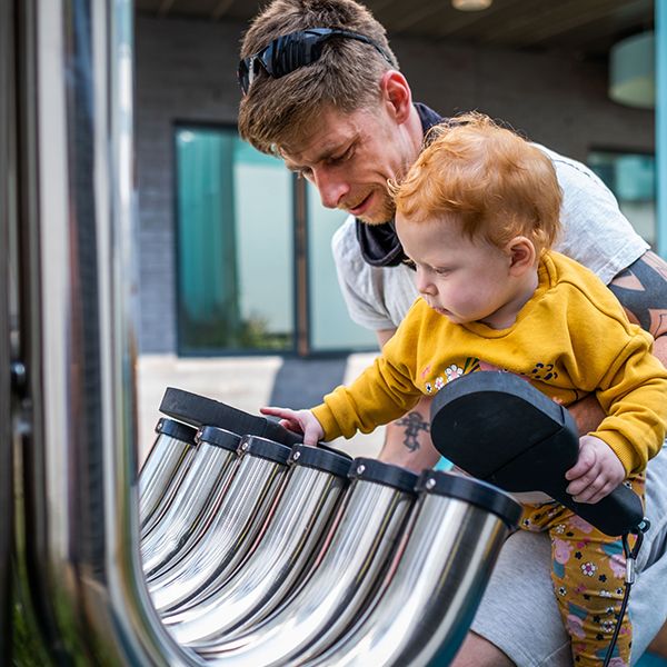 a young red headed girl and her father playing on large outdoor stainless steel aerophone tubes at the Edinburgh Children's Hospital garden