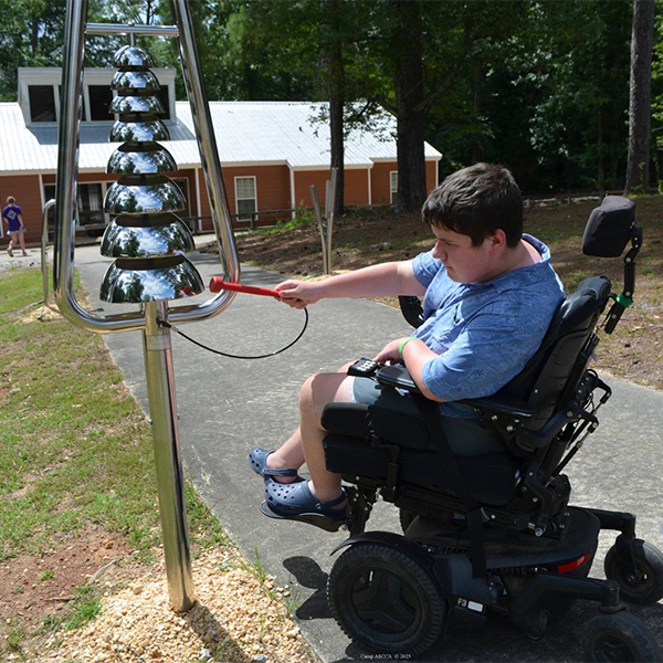 a male wheelchair user reaching up and playing a large stainless stell bell lyre outdoor musical instrument in a camp ground