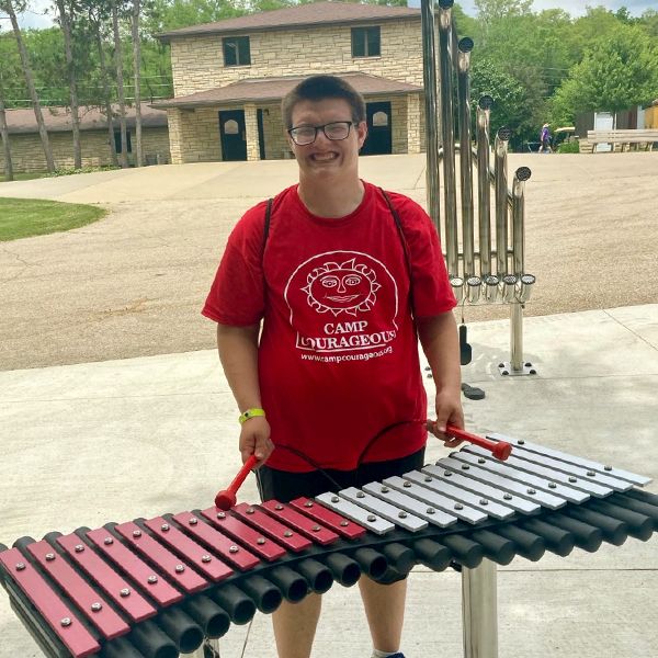 a young adult with special needs wearing a red t shirt and playing a large outdoor xylophone in a music park at summer camp 