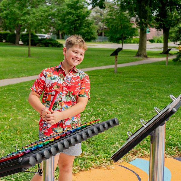 a boy in a floral shirt smiling while playing an outdoor xylophone in a music park