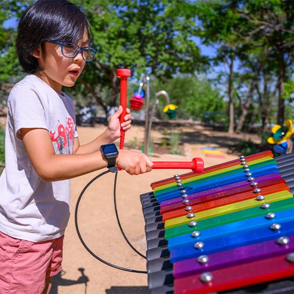 a young boy wearing thick rimmed glasses playing a colourful outdoor xylophone