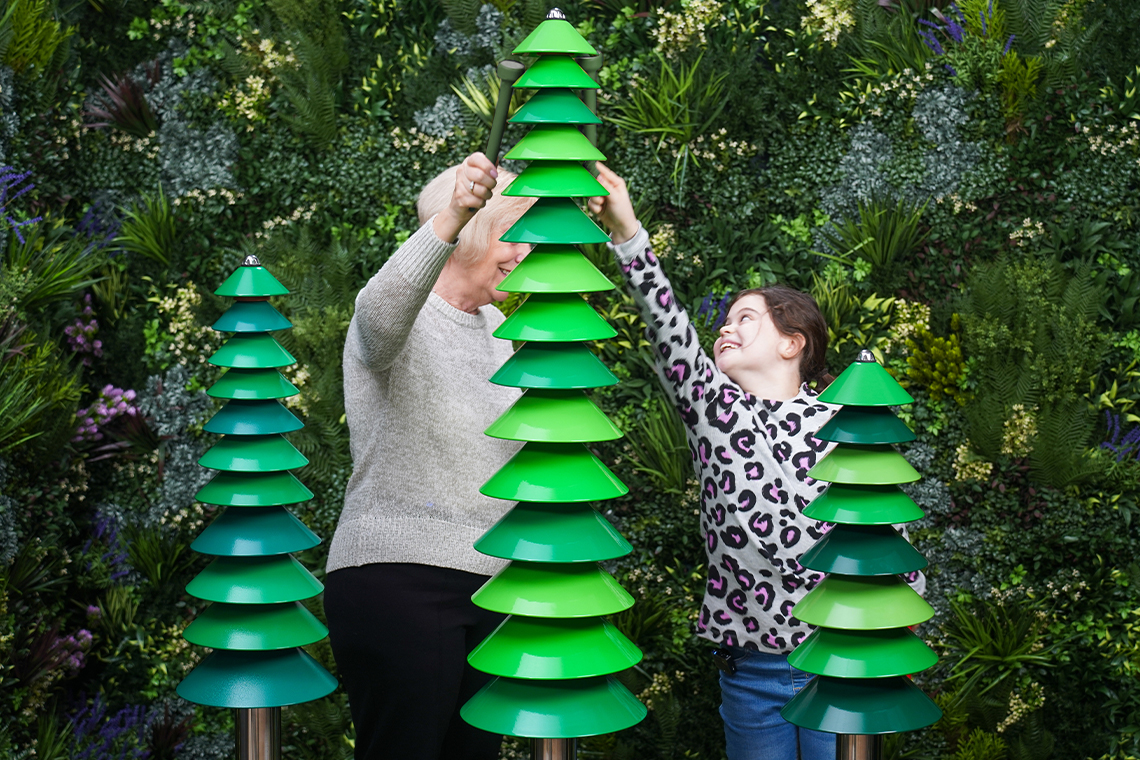 A woman and a child play green, tree-shaped musical bell structures resembling modern trees. They're smiling, set against a lush, leafy backdrop.