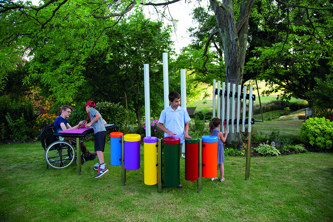 Children and a wheelchair user interact with colorful outdoor musical instruments in a park setting.