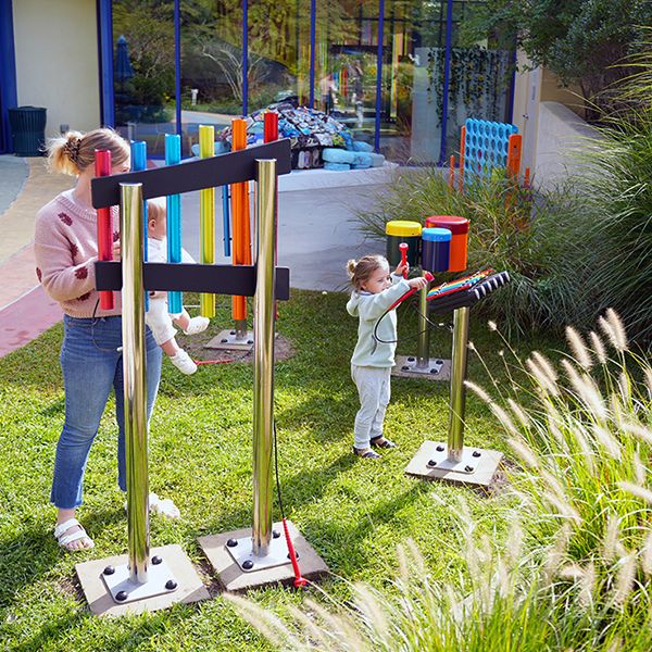 A young mother holding a bay playing outdoor musical instruments with another child in a museum sensory garden